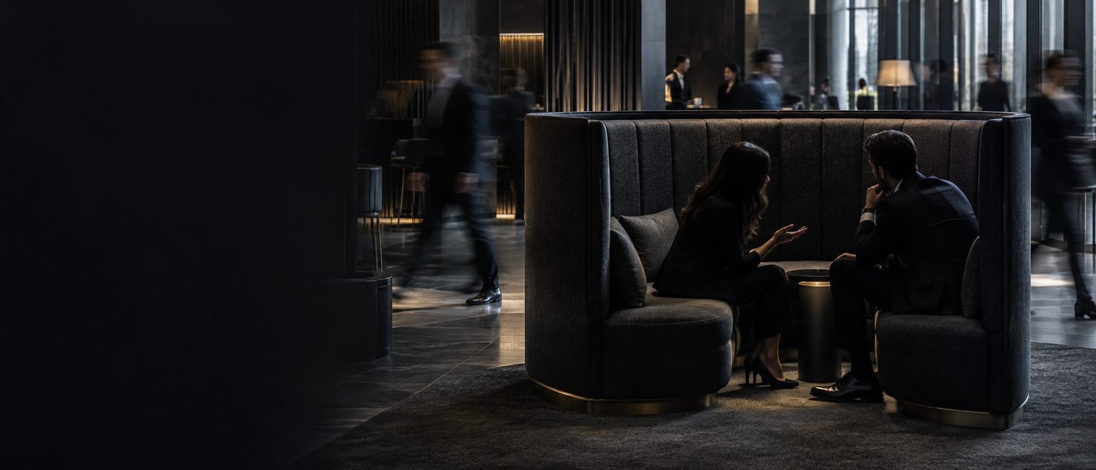 Two people in a dark, high-backed private booth within a busy contemporary space; the surrounding world continues in soft motion blur while their conversation stays calm, contained and separate.